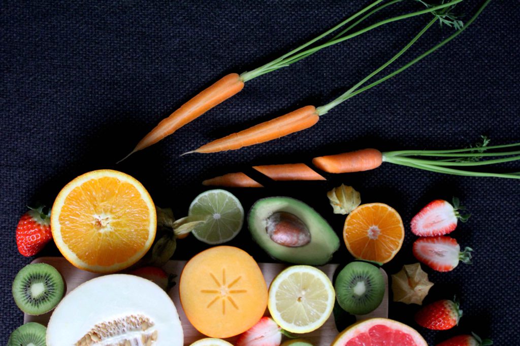 A selection of fruits and vegetables, including carrots, orange, melon, kiwi, avocado and strawberries against a black textured background.