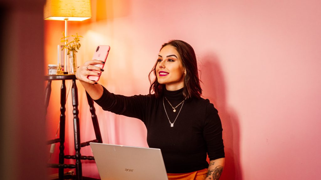 Female influencer with makeup on against a pink background taking a selfie.